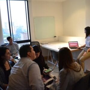 Un grupo de personas en una sala de reuniones, centrándose en una presentación de una mujer al frente junto a una pantalla digital y una pizarra.