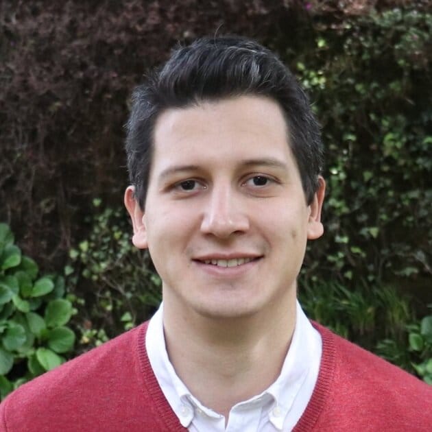 Un hombre con una leve sonrisa vestido con un suéter rojo sobre una camisa blanca, parado frente a arbustos de hojas verdes.