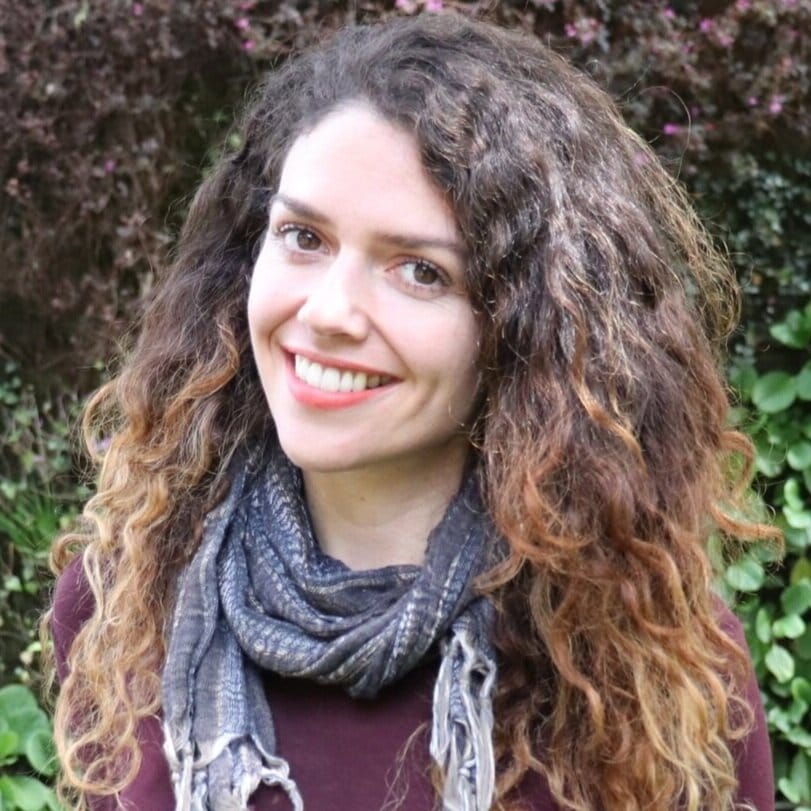 Una mujer sonriente con cabello rizado y una bufanda parada frente a un follaje verde.