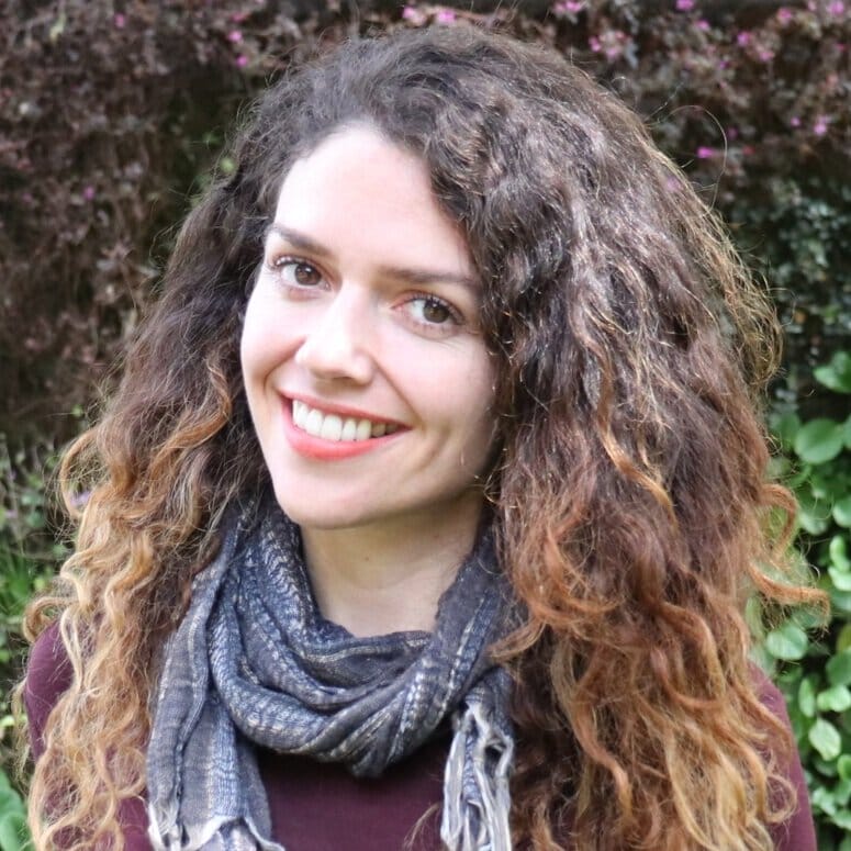 Una mujer sonriente con cabello castaño rizado y una bufanda gris, parada al aire libre con vegetación de fondo.