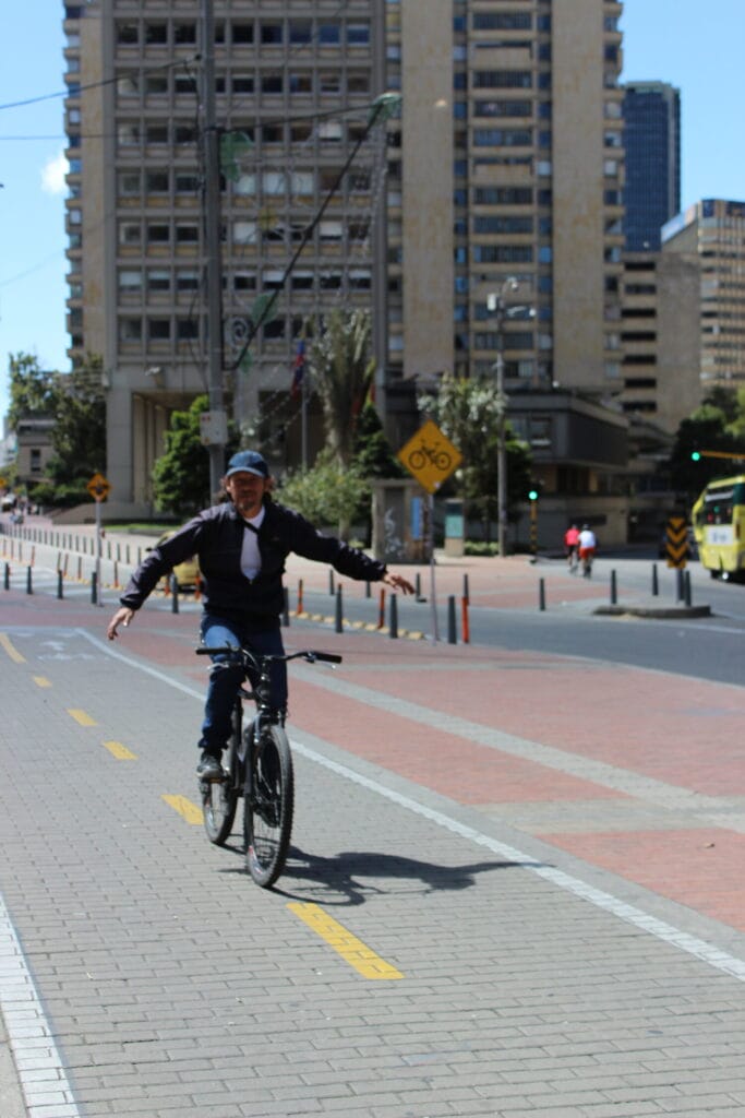 Un hombre andando en bicicleta sin manos en el manillar, sonriendo, en una calle de la ciudad con edificios altos al fondo.