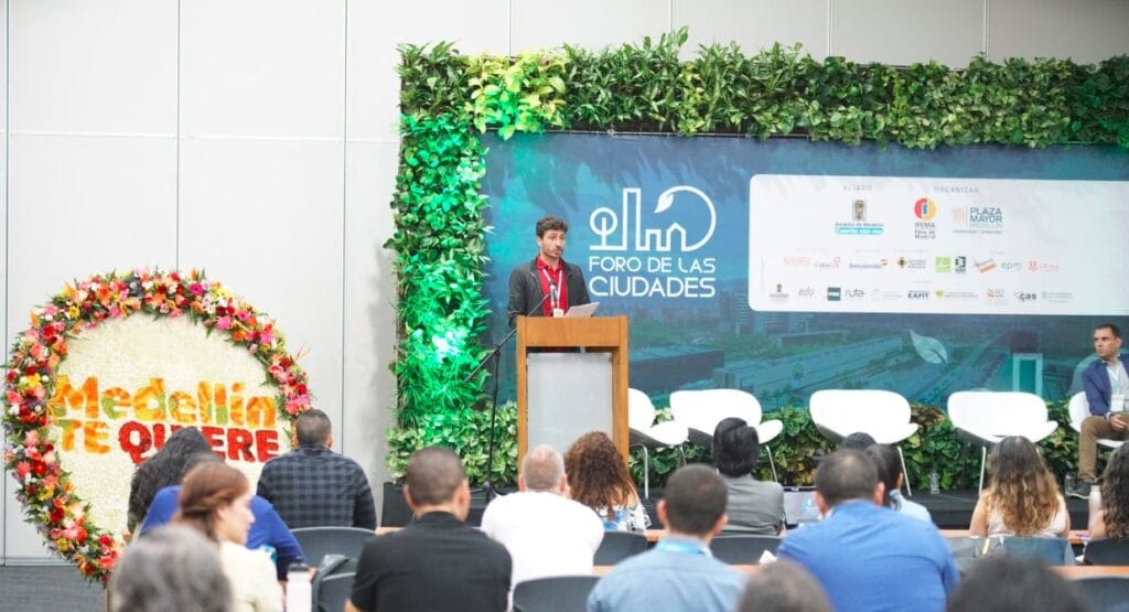 Un hombre habla en un podio durante una conferencia con un panel y una audiencia escuchando, y un fondo floral que dice "medellín te quiere".