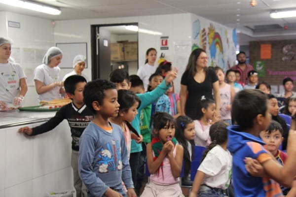 Niños de distintas edades haciendo cola en una cafetería, con adultos supervisando y una mujer sirviendo comida al fondo.