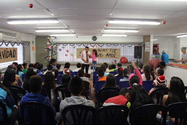 Una profesora dando una lección a un aula llena de niños, con adornos navideños y grandes pancartas en las paredes.