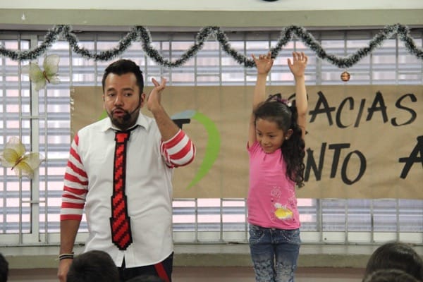 Un hombre y una joven interactuando alegremente en el escenario, con la niña levantando la mano, en una sala decorada con guirnaldas.