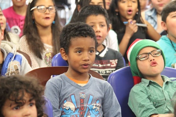 Grupo de niños diversos sentados y mirando atentamente; un niño con un sombrero verde parece estar bostezando.