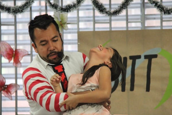 Un hombre con una camisa a rayas sostiene juguetonamente a una joven riendo en un evento festivo con decoraciones en el fondo.