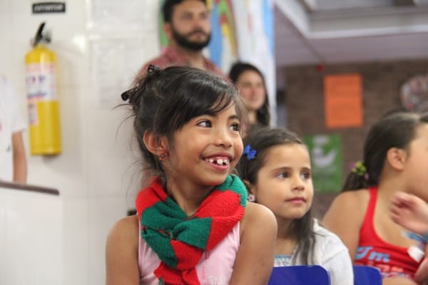 Niña sonriendo en un evento, luciendo un pañuelo rojo y verde, con otras personas al fondo.