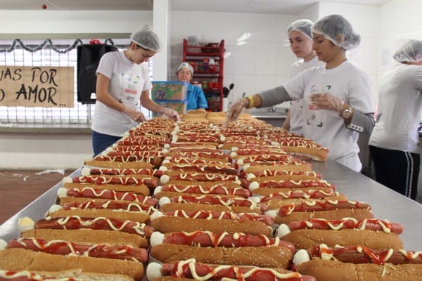 Voluntarios con redecillas para el pelo preparan perritos calientes en una mesa larga en una cocina comunitaria.