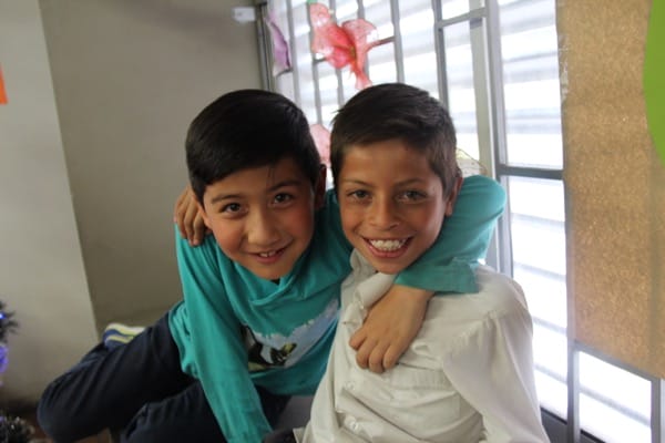 Dos niños sonrientes abrazados, sentados en el interior con decoraciones en el fondo.