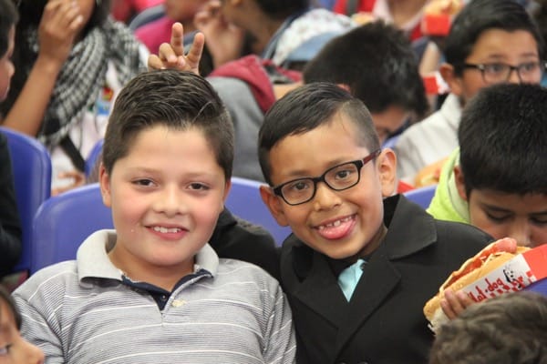 Dos niños sonriendo a la cámara, uno de ellos haciendo un signo de la paz, rodeados de otros niños en una habitación llena de gente.