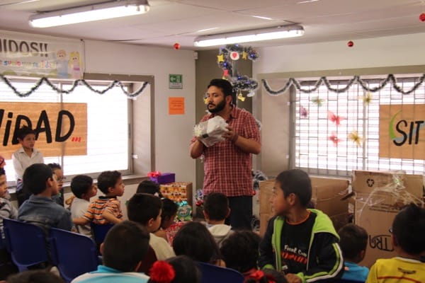 Un hombre que sostiene un conejo habla con niños sentados en un salón de clases decorado con guirnaldas festivas y recortes de papel.