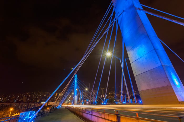 Vista nocturna de un puente atirantado iluminado por luces azules con el horizonte de la ciudad al fondo.