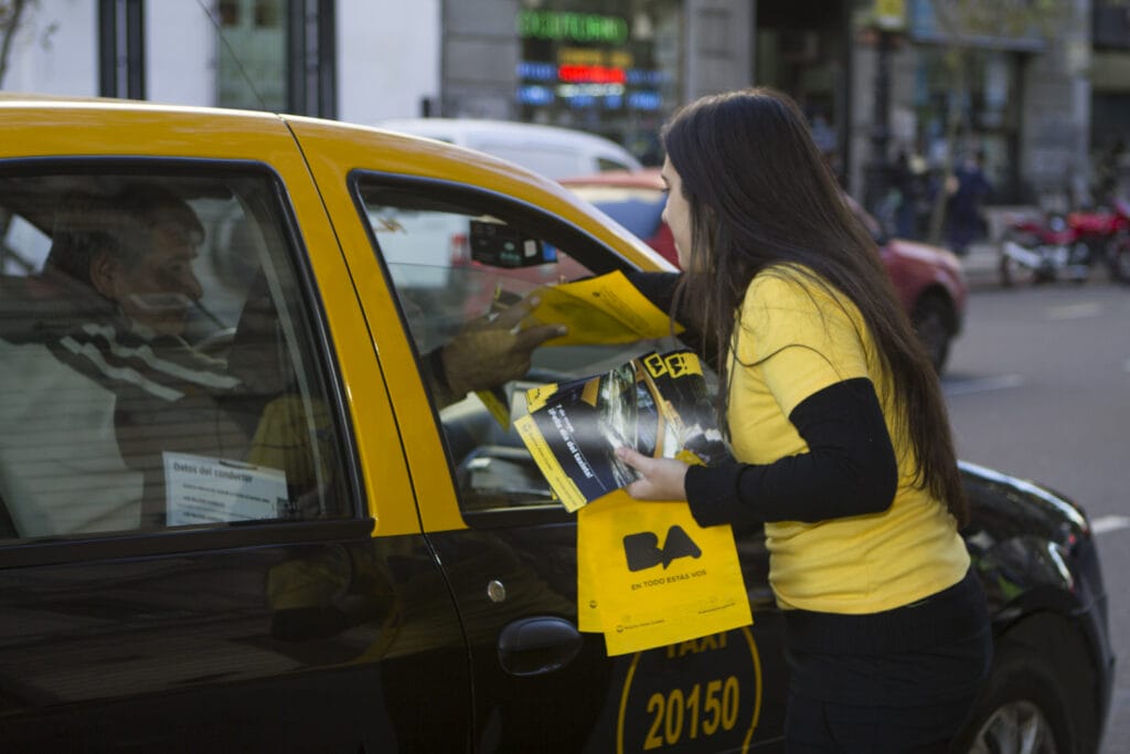 Una mujer con una camisa amarilla reparte folletos a un hombre en un taxi amarillo en una calle de la ciudad.