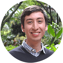 Un joven con una leve sonrisa, vestido con una camisa a cuadros debajo de un suéter, posando frente a un exuberante follaje verde.