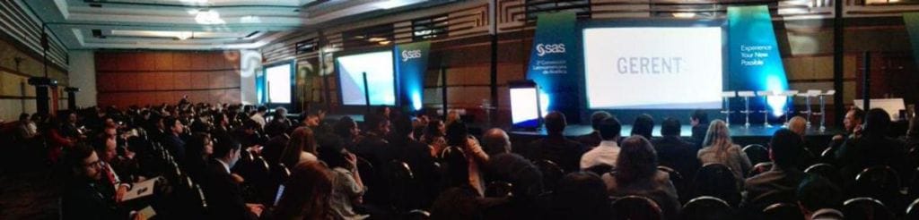 Vista gran angular de una conferencia de negocios con los asistentes sentados frente a dos grandes pantallas de proyección que muestran logotipos y texto.