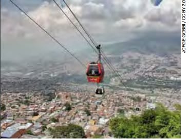 Teleféricos rojos que ascienden sobre una ciudad en expansión con montañas distantes y un cielo nublado al fondo.