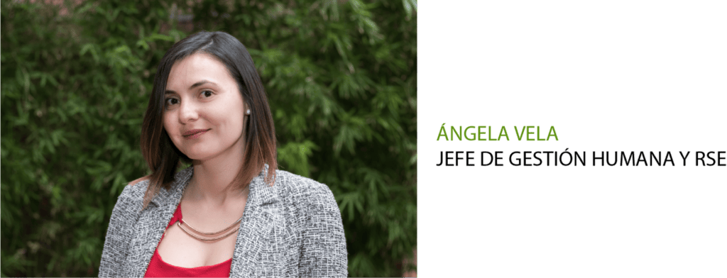 Mujer con top rojo y chaqueta gris sonriendo, de pie sobre un fondo verde con el texto "ángela vela" sobre fondo negro.