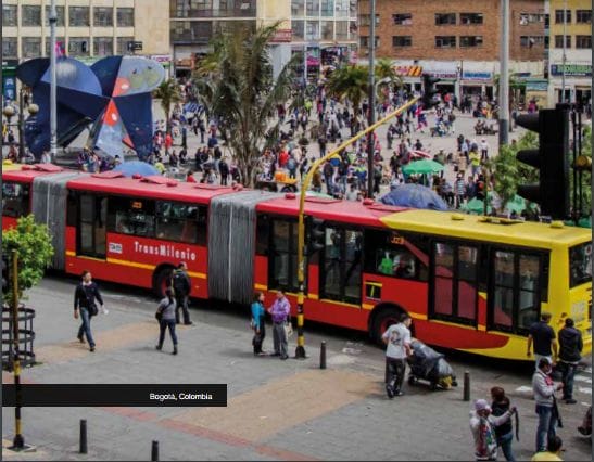 Una animada escena urbana en bogotá, colombia, con una multitud de personas, un autobús transmilenio rojo y amarillo y edificios modernos bajo un cielo nublado.