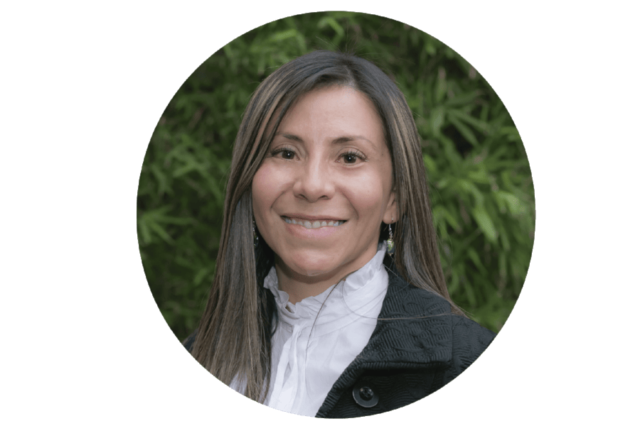 Retrato de una mujer sonriente con el pelo largo, vestida con una chaqueta negra y una blusa blanca, con un fondo de bambú.
