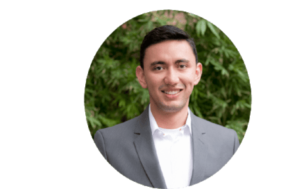 Una foto profesional de la cabeza de un hombre sonriente con traje gris y camisa blanca, de pie frente a un fondo de follaje verde borroso.