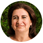 Retrato de una mujer sonriente con cabello castaño hasta los hombros y aretes, sobre un fondo de follaje verde borroso.