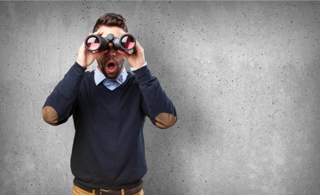 Un hombre con una expresión de sorpresa mirando a través de binoculares sobre un fondo gris texturizado.
