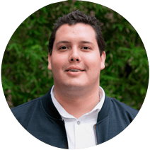 Un retrato de un hombre sonriente con una chaqueta negra y una camisa blanca sobre un fondo de follaje verde.
