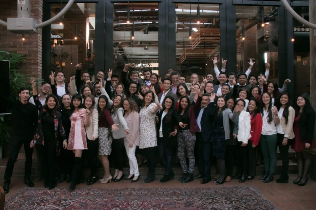 Un gran grupo de personas posando para una fotografía en un evento bajo techo, sonriendo y levantando la mano con alegría.