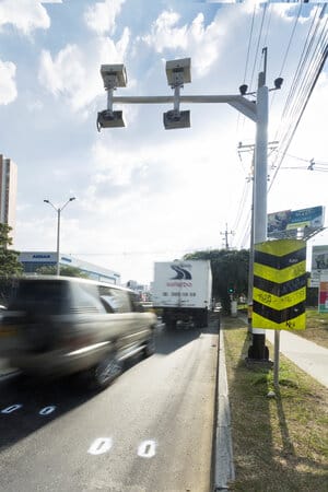 Cámaras de vigilancia del tráfico en un poste sobre una carretera muy transitada con vehículos a alta velocidad, bajo un cielo despejado.