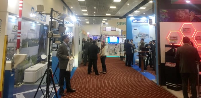 Piso de feria con varios stands de tecnología que muestran equipos y varios asistentes conversando.