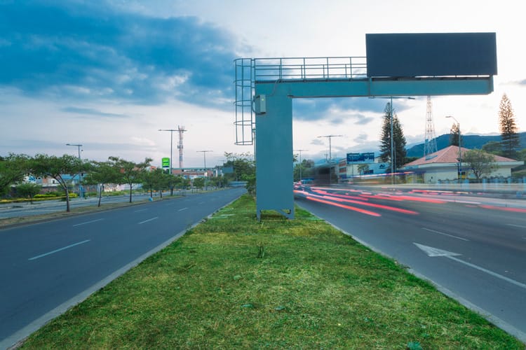 Una carretera con estelas de luz al anochecer, con un gran cartel en blanco y un fondo montañoso.