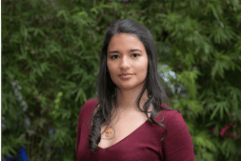 Una mujer joven con el pelo largo y oscuro recogido en una trenza, vestida con un top granate, parada frente a un follaje verde.