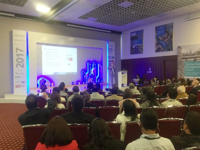 Un orador presenta una conferencia frente a una audiencia, con una pantalla grande que muestra una diapositiva de PowerPoint.