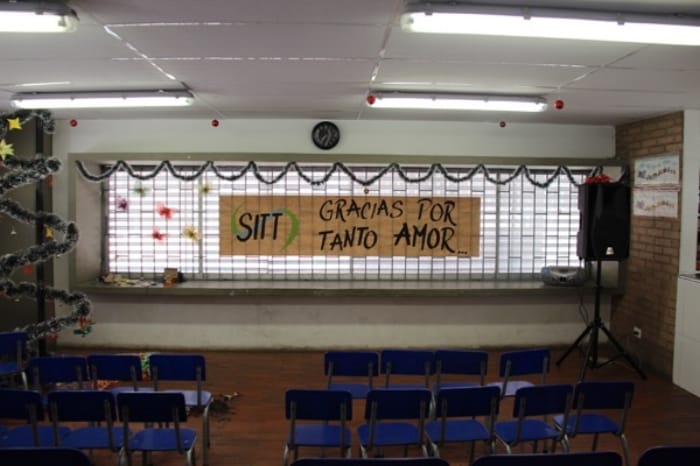 Aula decorada con adornos navideños, una pancarta que dice "gracias por tanto amor", sillas vacías y un altavoz.