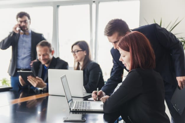 Grupo de profesionales en una reunión, con un hombre y una mujer trabajando en una computadora portátil mientras otros discuten y usan tabletas digitales en una oficina moderna.