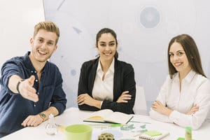 Tres colegas en una mesa de reuniones, un hombre extendiendo un apretón de manos y dos mujeres sonriendo a la cámara, en un ambiente de oficina luminoso. Tres colegas en una mesa de reuniones, un hombre extendiendo un apretón de manos y dos mujeres sonriendo a la cámara, en un ambiente de oficina luminoso.