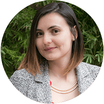 Una mujer con cabello castaño hasta los hombros y vestida con una chaqueta gris sonríe sobre un fondo de follaje verde.