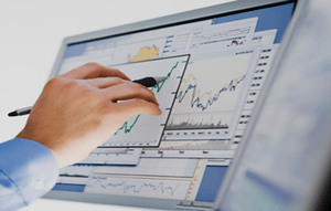 Primer plano de la mano de una persona sosteniendo un bolígrafo, apuntando a gráficos financieros en la pantalla de una computadora.