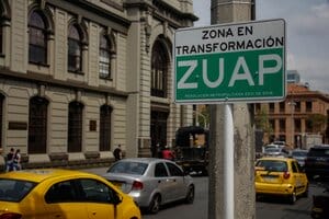Letrero que dice "zona en transformación zuap" frente a una calle muy transitada con autos que pasan y un edificio clásico al fondo, designado como parte de "Las Zonas Urbanas de