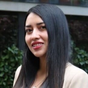 Una mujer con cabello largo y negro y una chaqueta beige sonriendo al aire libre.