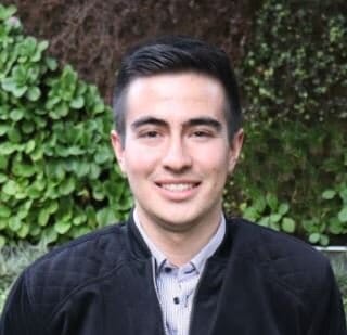 Un joven sonriente con cabello corto y oscuro, vestido con una chaqueta negra sobre una camisa gris, posando frente a un seto de hojas verdes.