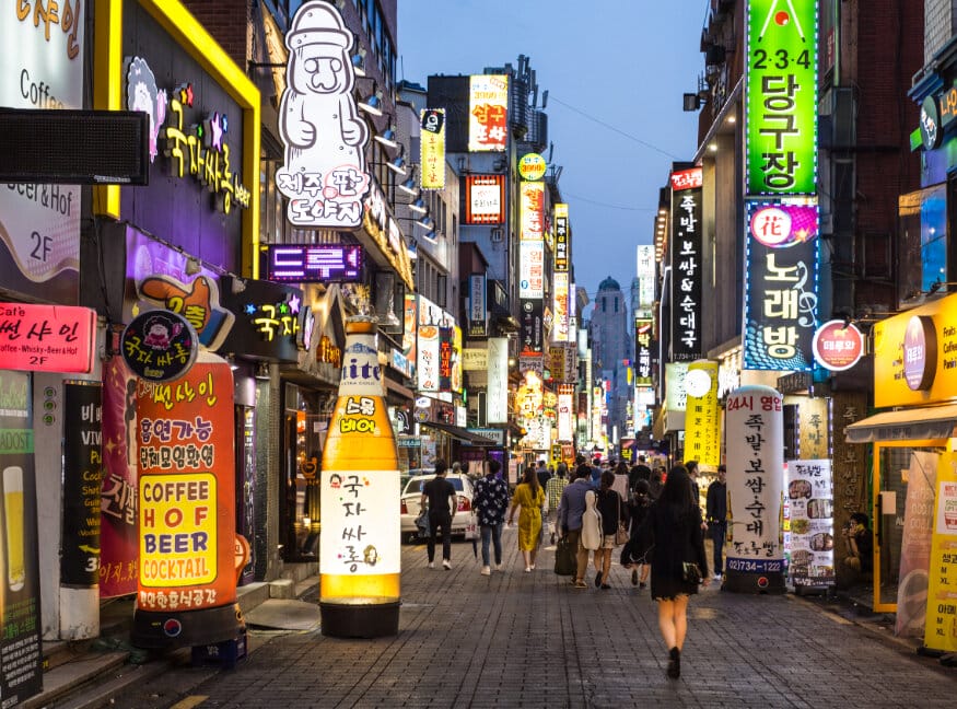 Calle brillantemente iluminada en Corea del Sur por la noche, llena de gente y llena de carteles de neón que anuncian tiendas, cafeterías y bares.