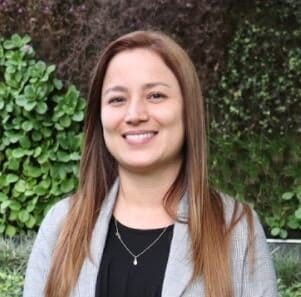 Una mujer sonriente con cabello castaño hasta los hombros, vestida con una chaqueta gris, se encuentra frente a un seto de hojas verdes.