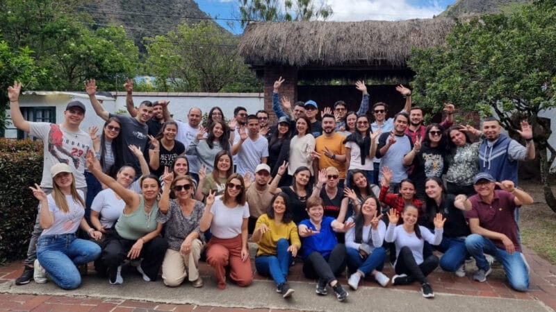 Un gran grupo de personas, sonriendo y levantando las manos, posan al aire libre frente a un edificio con techo de paja y un paisaje montañoso al fondo.