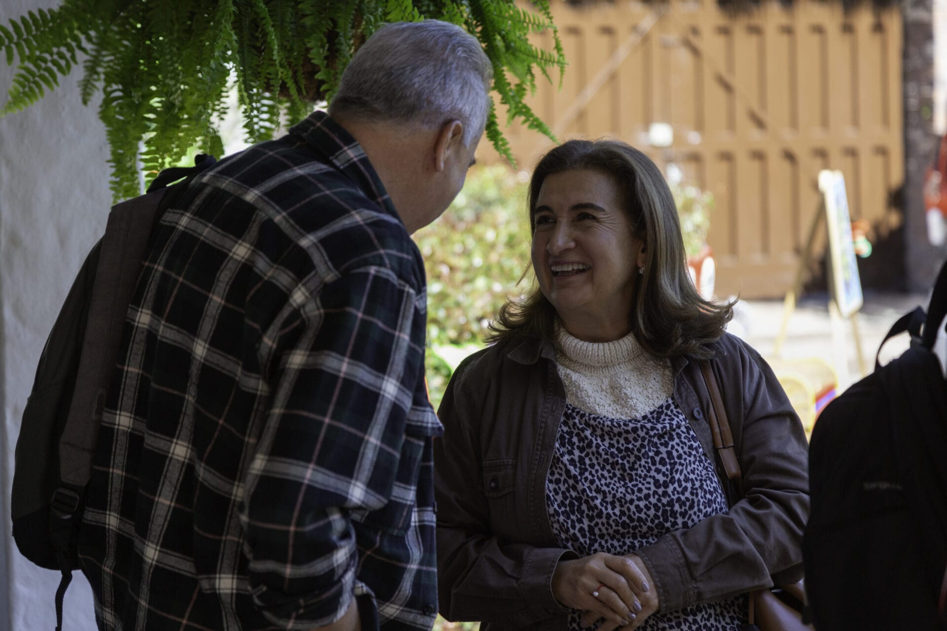 Un hombre y una mujer conversan al aire libre cerca de un helecho, con la mujer sonriendo y juntando las manos.