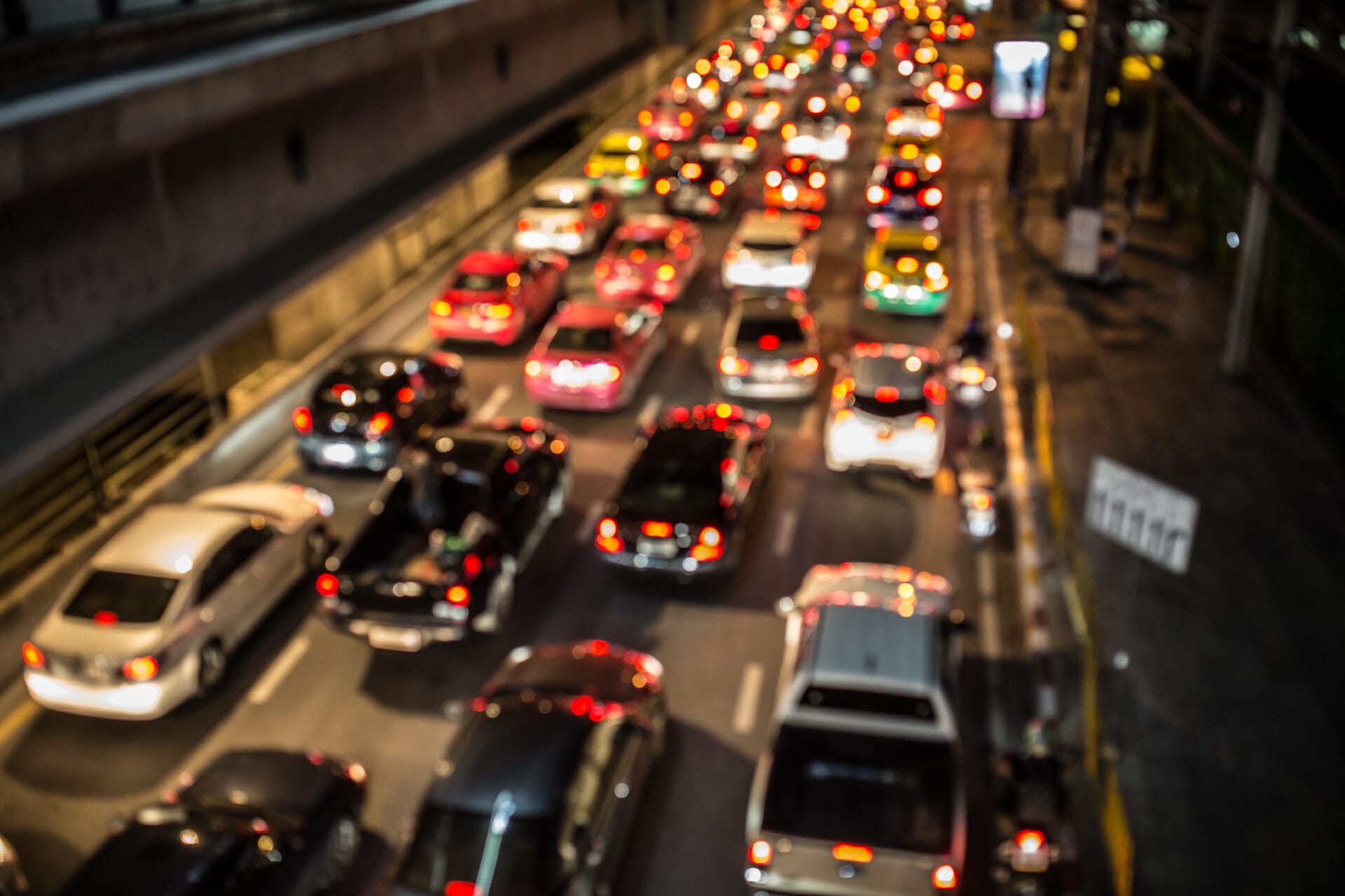 Descripción: Imagen borrosa de tráfico en una ciudad sostenible por la noche mostrando múltiples autos con faros y luces traseras encendidas en una calle concurrida.