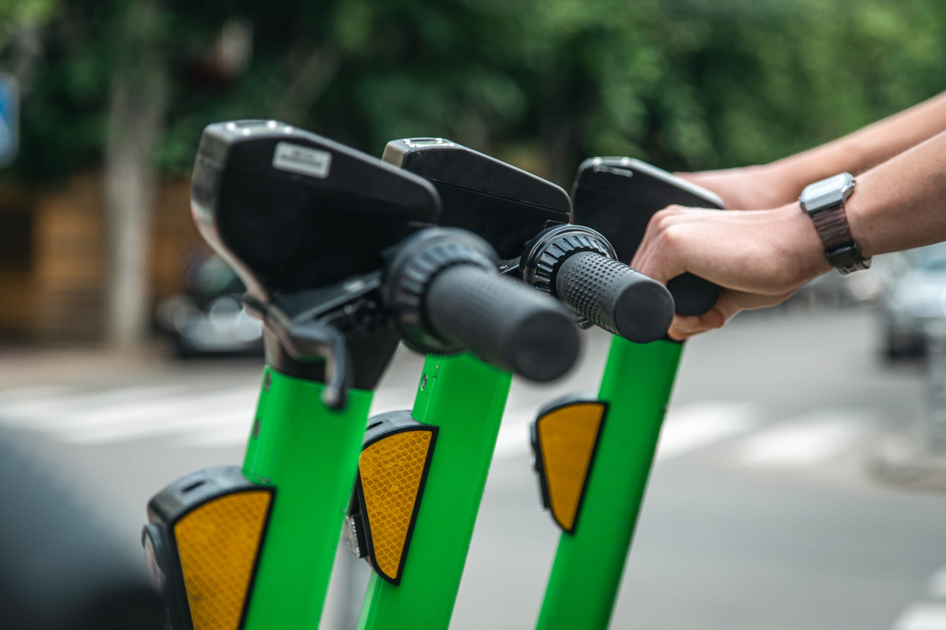 Primer plano de manos sosteniendo el manillar de dos scooters eléctricos verdes en una calle de la ciudad, centrándose en opciones de movilidad limpia.