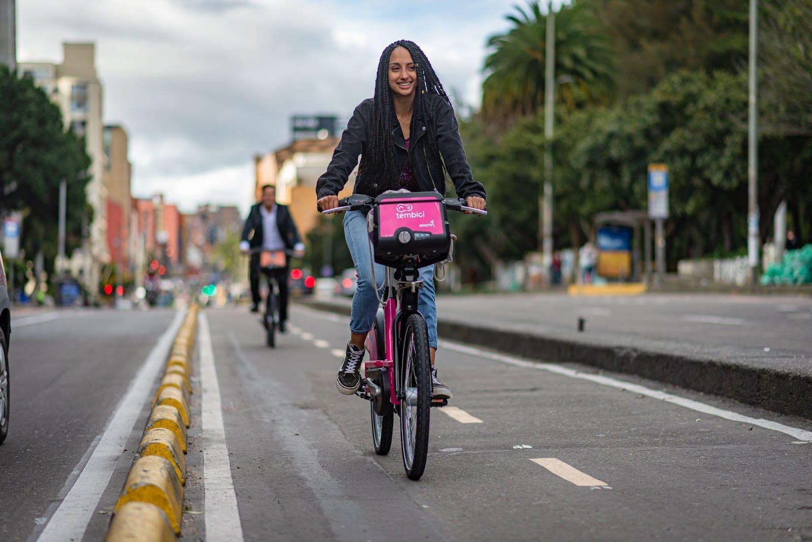 Una joven sonriendo mientras anda en bicicleta por una calle de la ciudad, con una canasta rosa en su bicicleta y un fondo borroso de otros ciclistas y vegetación, promoviendo la movilidad sostenible.