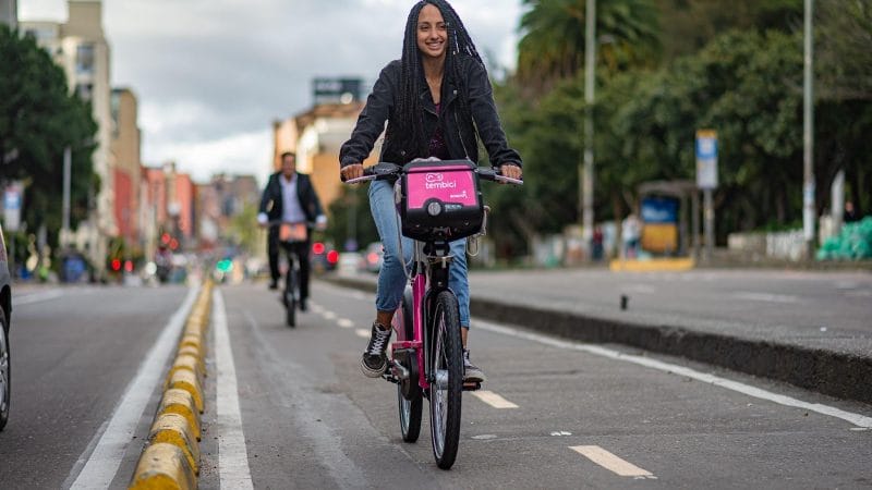 Una joven sonriendo mientras anda en bicicleta por una calle de la ciudad, con una canasta rosa en su bicicleta y un fondo borroso de otros ciclistas y vegetación, promoviendo la movilidad sostenible.
